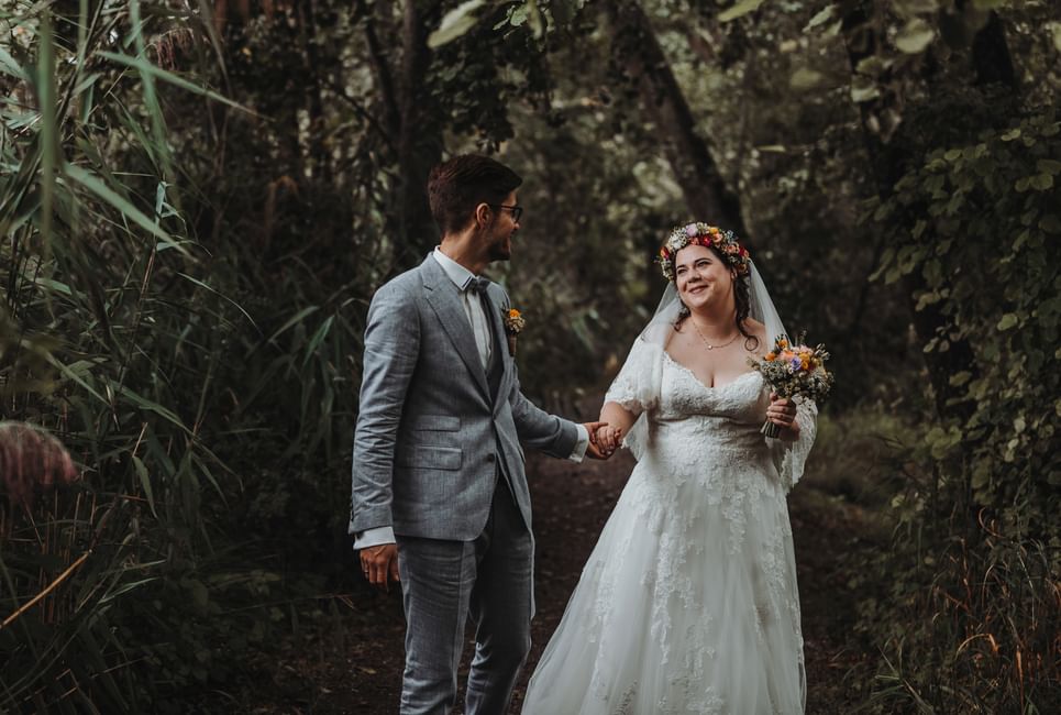 Ein Brautpaar auf einem Pfad im Wald. Der Bräutigam führt die Braut an der Hand. Die Braut hält einen Blumenstrauss.